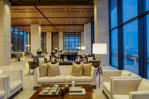 A spacious lobby with a modern design, featuring a seating area with sofas and armchairs, a dining area with tables, and large windows offering views.