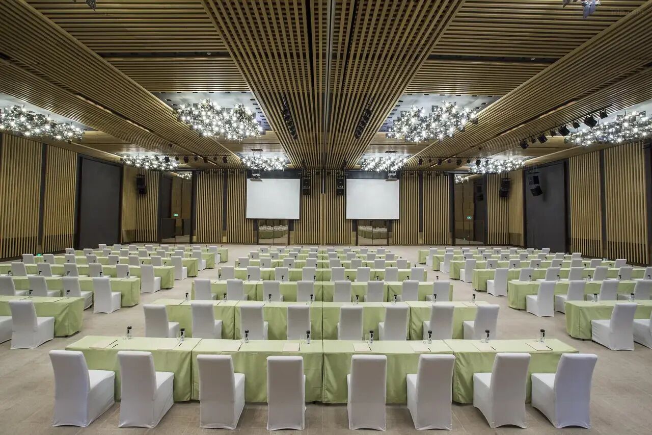 A conference room with rows of tables and chairs, a high ceiling with a wooden slat design, and large screens at the front.