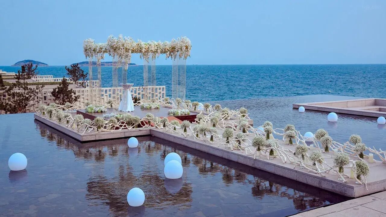 A pool area with a view of the sea, a covered seating area, and decorative plants.
