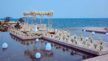 A pool area with a view of the sea, a covered seating area, and decorative plants.