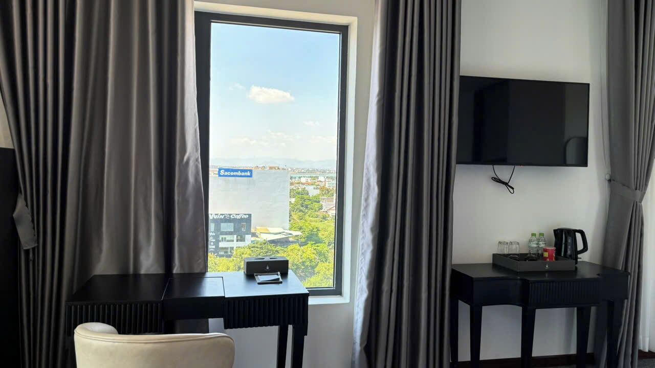 A hotel room with a large window, a flat-screen TV, a desk with a kettle, and a chair.