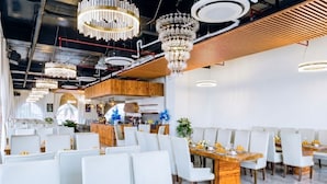 Interior moderno de restaurante con varias mesas, sillas blancas y una variedad de luminarias, incluyendo candelabros y lámparas colgantes.