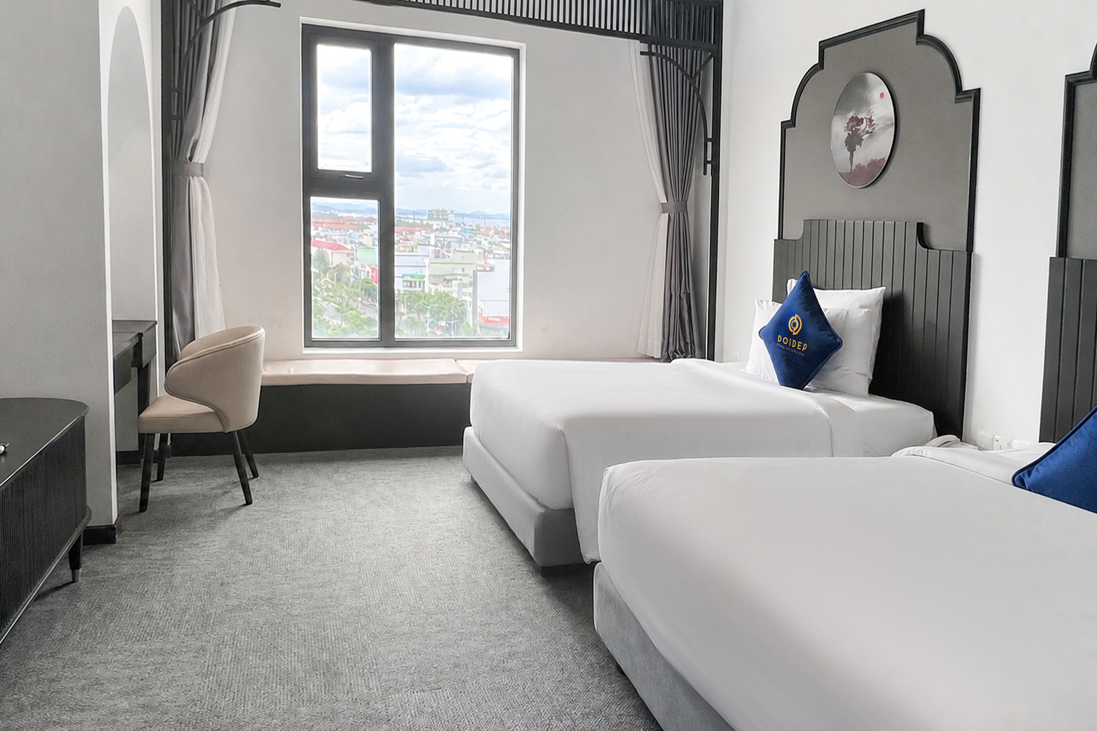 Habitación de hotel con dos camas, un escritorio, una silla, un ventanal grande con vista a la ciudad y un elemento decorativo en la pared.