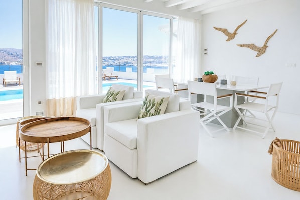 Una sala de estar moderna con vista al mar, muebles blancos y una zona de comedor.