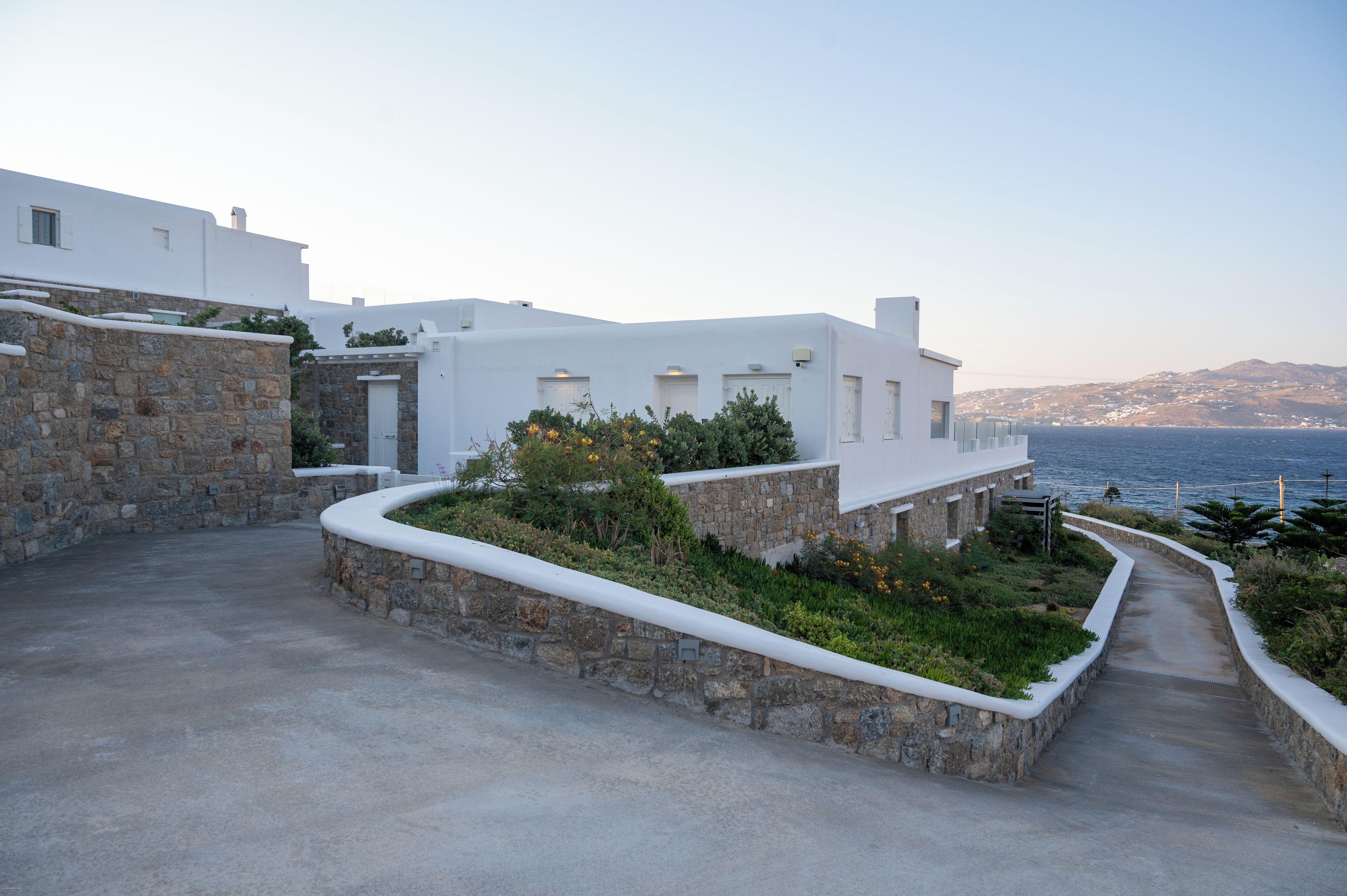 Un edificio costero con paredes blancas y cimientos de piedra, un camino empedrado y vistas al mar y colinas lejanas.