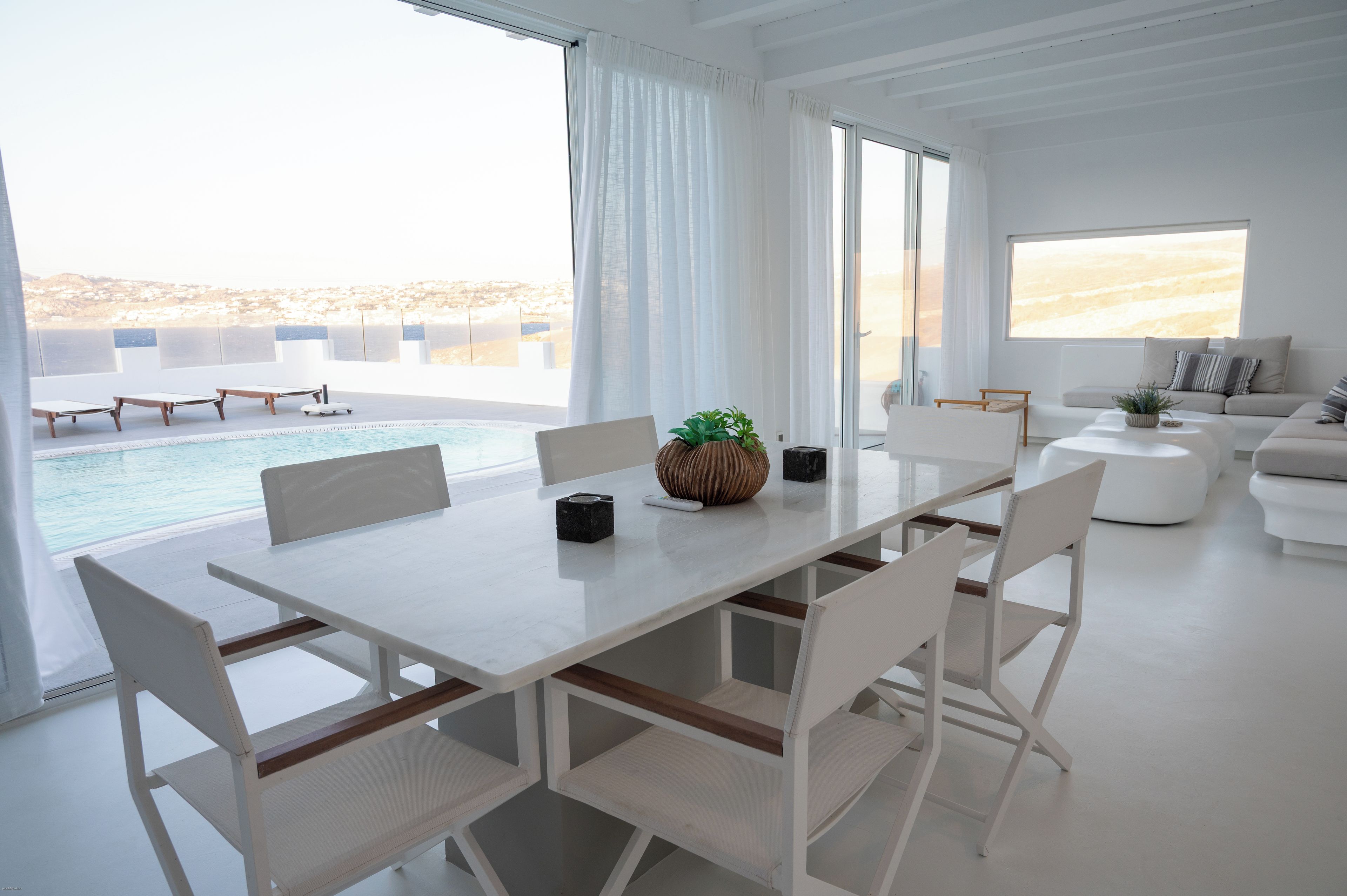 Sala de estar moderna com uma mesa grande branca, cadeiras brancas, um sofá e vista para uma piscina e praia.