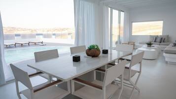 Sala de estar moderna com uma mesa grande branca, cadeiras brancas, um sofá e vista para uma piscina e praia.