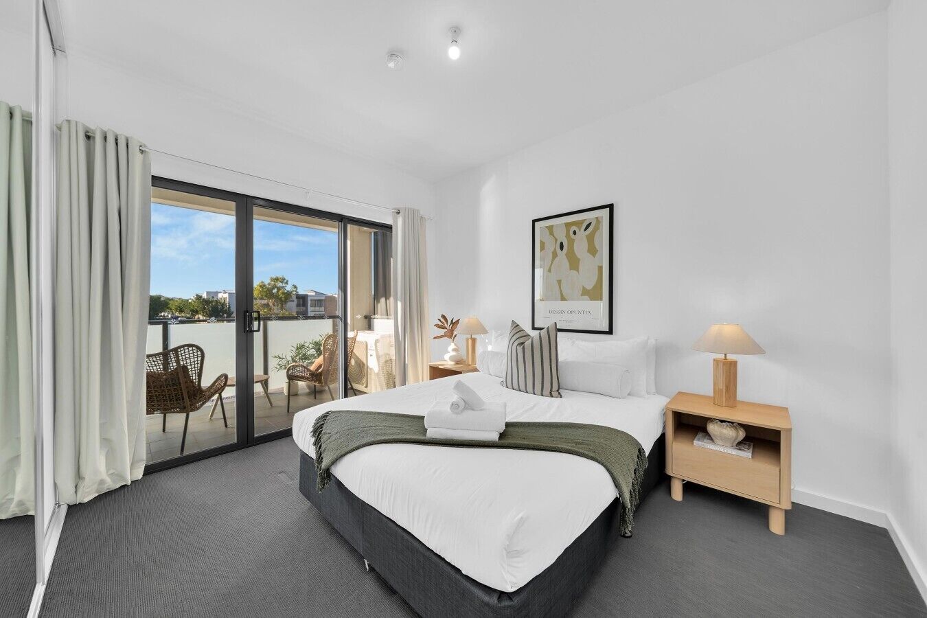 A hotel room with a bed, a nightstand, a lamp, a framed picture, and a view of the outdoors through a sliding glass door.