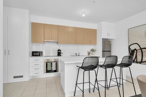 A modern kitchen with white cabinets, a stainless steel oven, a microwave, and a coffee maker. There are three black chairs with a unique design. - Lakeside Nook | By Host Solutions (Lightsview)