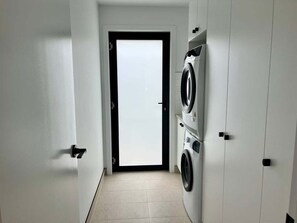 A laundry room with a washing machine and dryer, a tiled floor, and a door leading to another room. - Ocean Mist Cabin – Spa, Fireplace & EV Charger (Huon Valley)