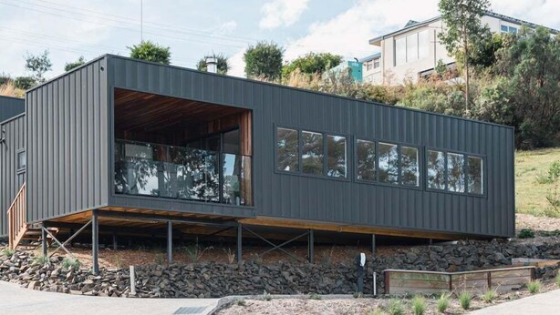 A modern building with a flat roof, large windows, and a wooden deck area. - Ocean Mist Cabin – Spa, Fireplace & EV Charger (Huon Valley)