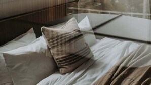 A bed with white bedding and a brown throw blanket, a glass table, and a view of a lake through a window. - Ocean Mist Cabin – Spa, Fireplace & EV Charger (Huon Valley)