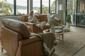 A modern living room with a leather sofa, a coffee table, and a view of the outdoors. - Ocean Mist Cabin – Spa, Fireplace & EV Charger (Huon Valley)