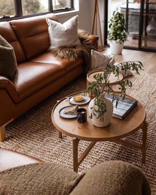 A cozy living room with a brown leather sofa, a round wooden coffee table with a plant, a book, and a mug.