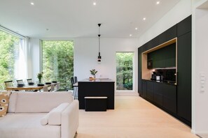A modern kitchen with a black countertop, a dining area with a white sofa, and a view of greenery through large windows. - Christmas Villa, Private Sauna By Lapland Host (Rovaniemi)