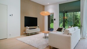 Ein modernes Wohnzimmer mit Flachbildfernseher, einem weißen Sofa, einem weißen Teppich und einem großen Fenster mit Blick auf Grünflächen.