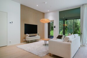 A modern living room with a flat-screen TV, a white sofa, a white rug, and a large window offering a view of greenery. - Christmas Villa, Private Sauna By Lapland Host (Rovaniemi)