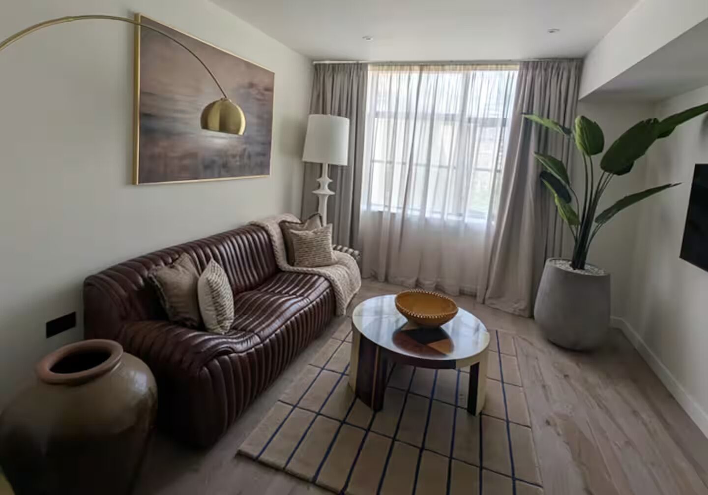 A living room with a brown leather sofa, a round coffee table, a large potted plant, and a painting on the wall.