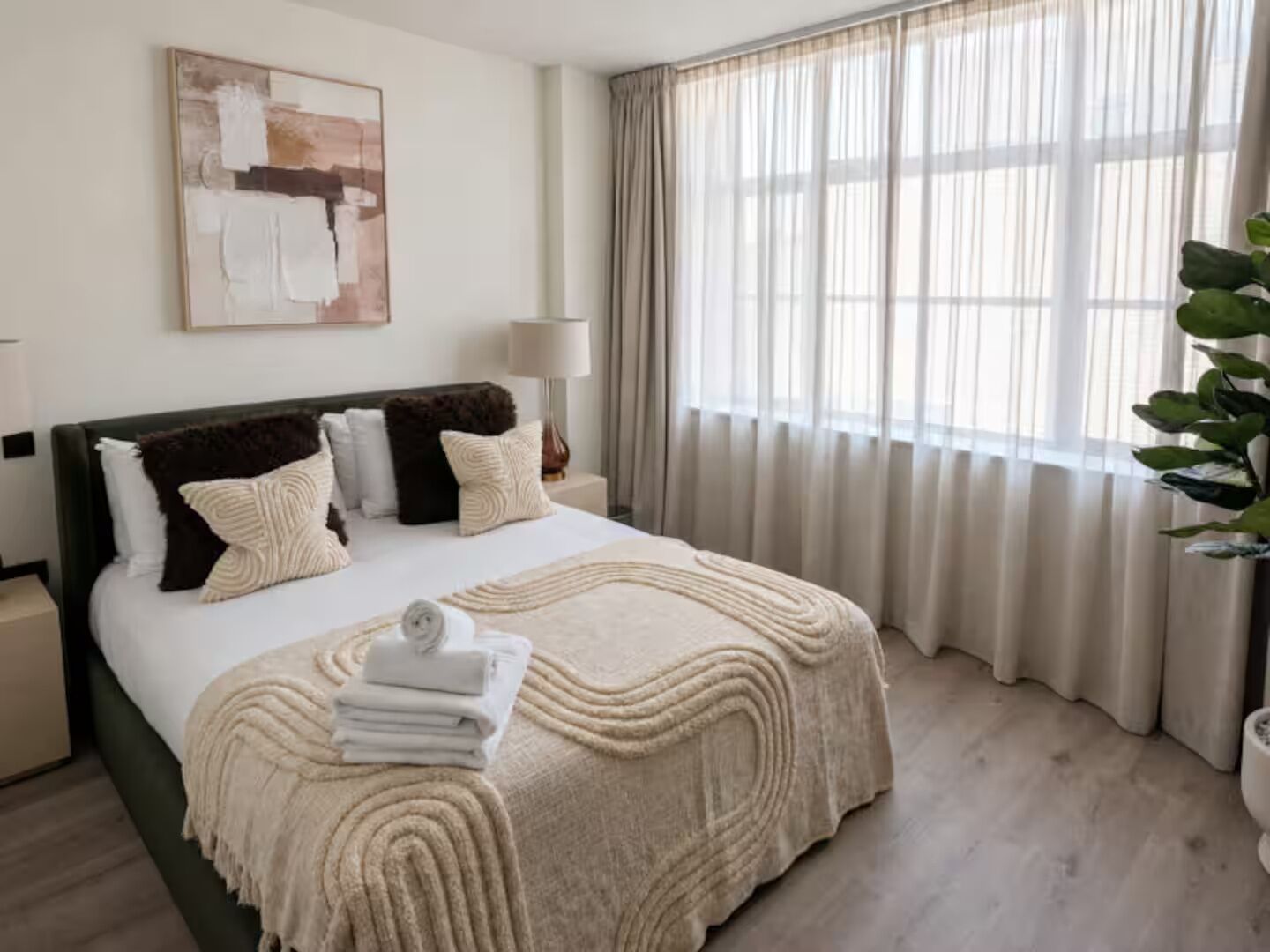 Ein Hotelzimmer mit Bett, Nachttisch, Lampe, Gemälde und einem Fenster mit Vorhängen.