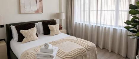 Una habitación de hotel con una cama, una mesita de noche, una lámpara, un cuadro y una ventana con cortinas.