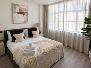 Een hotelkamer met een bed, nachtkastje, lamp, schilderij en raam met gordijnen.