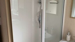 A glass-enclosed shower stall with a handheld showerhead, a white sink with a mirror above it, and a small shelf with toiletries. - Dinas Cross Country Park - Holiday Accommodation 20053 (Newport)