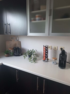 A kitchen with dark cabinets, a white countertop, a coffee maker, a plant, a mug rack, and a water dispenser. - Vacation Rental - Single-Family Home (Saint-Louis)
