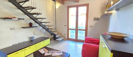 A living room with a staircase, a kitchen area, and a door leading outside.