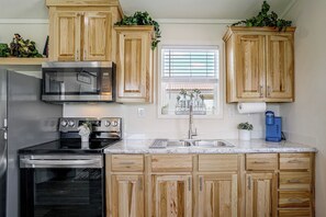 A kitchen with wooden cabinets, stainless steel appliances, a window with blinds, and a blue water dispenser. - Tiny Home Retreat Close to Lake Granbury (Granbury)