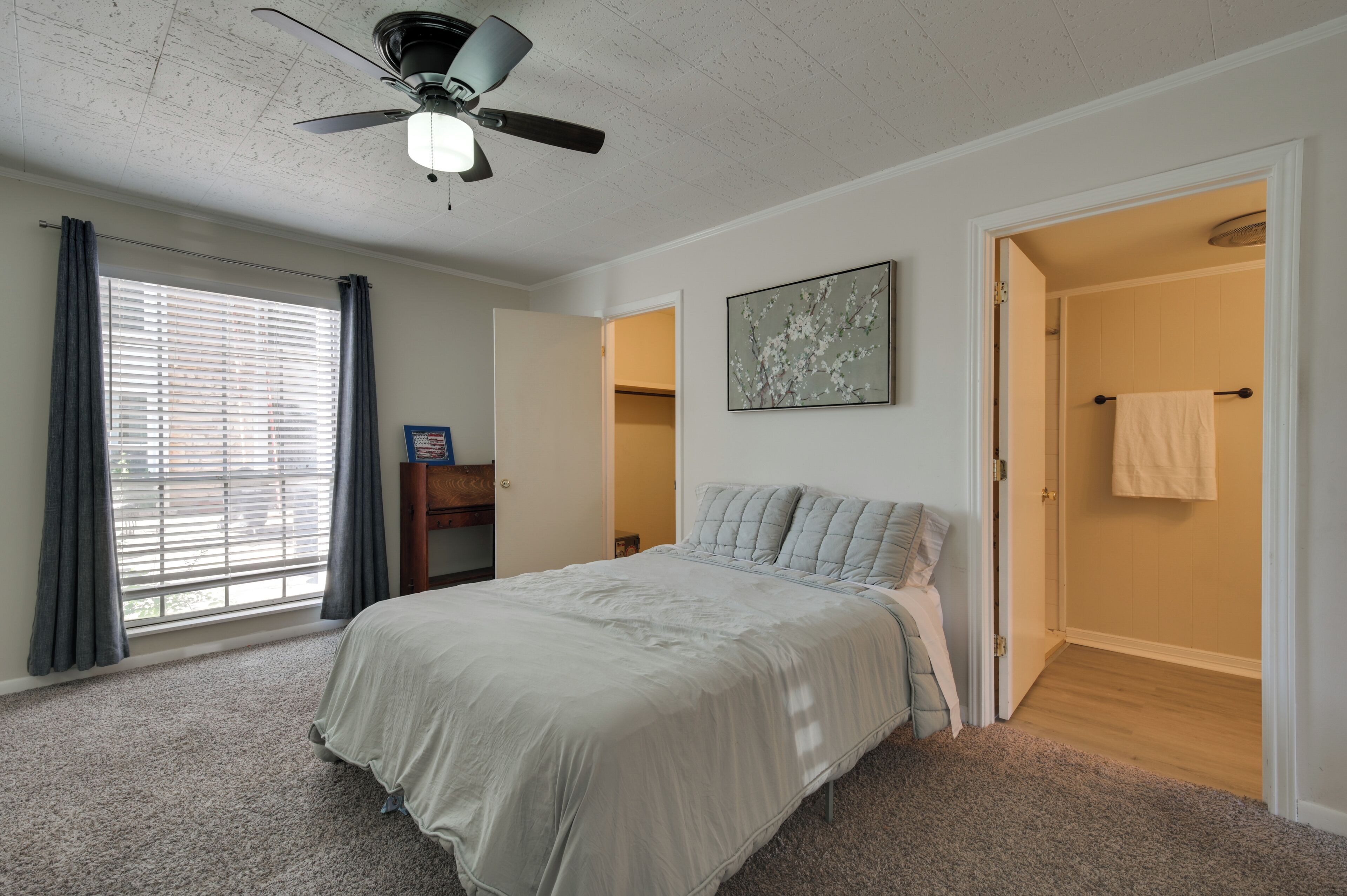 Une chambre à coucher avec un lit, un ventilateur de plafond, une fenêtre avec des rideaux, une porte donnant sur une salle de bain et un tableau accroché au mur.
