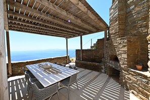 An outdoor patio with a table and chairs, a pergola, stone walls, and a view of the sea. - Dioni Tinos sea View Elegant Villa (Tinos)