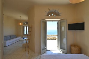 A bedroom with a bed, a television, a sofa, and a view of the sea through an open door. - Dioni Tinos sea View Elegant Villa (Tinos)