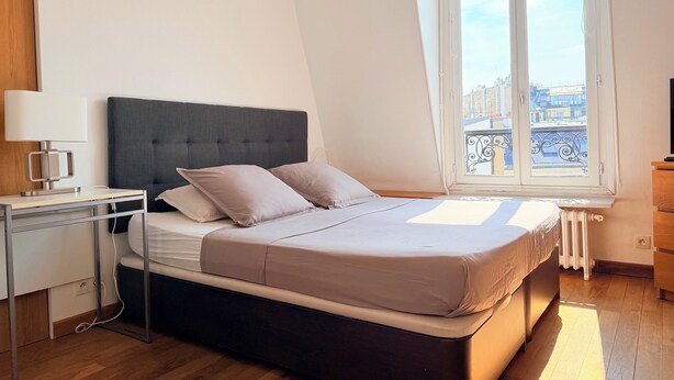 Un dormitorio con cama, mesita de noche, lámpara y ventana con vistas a edificios.
