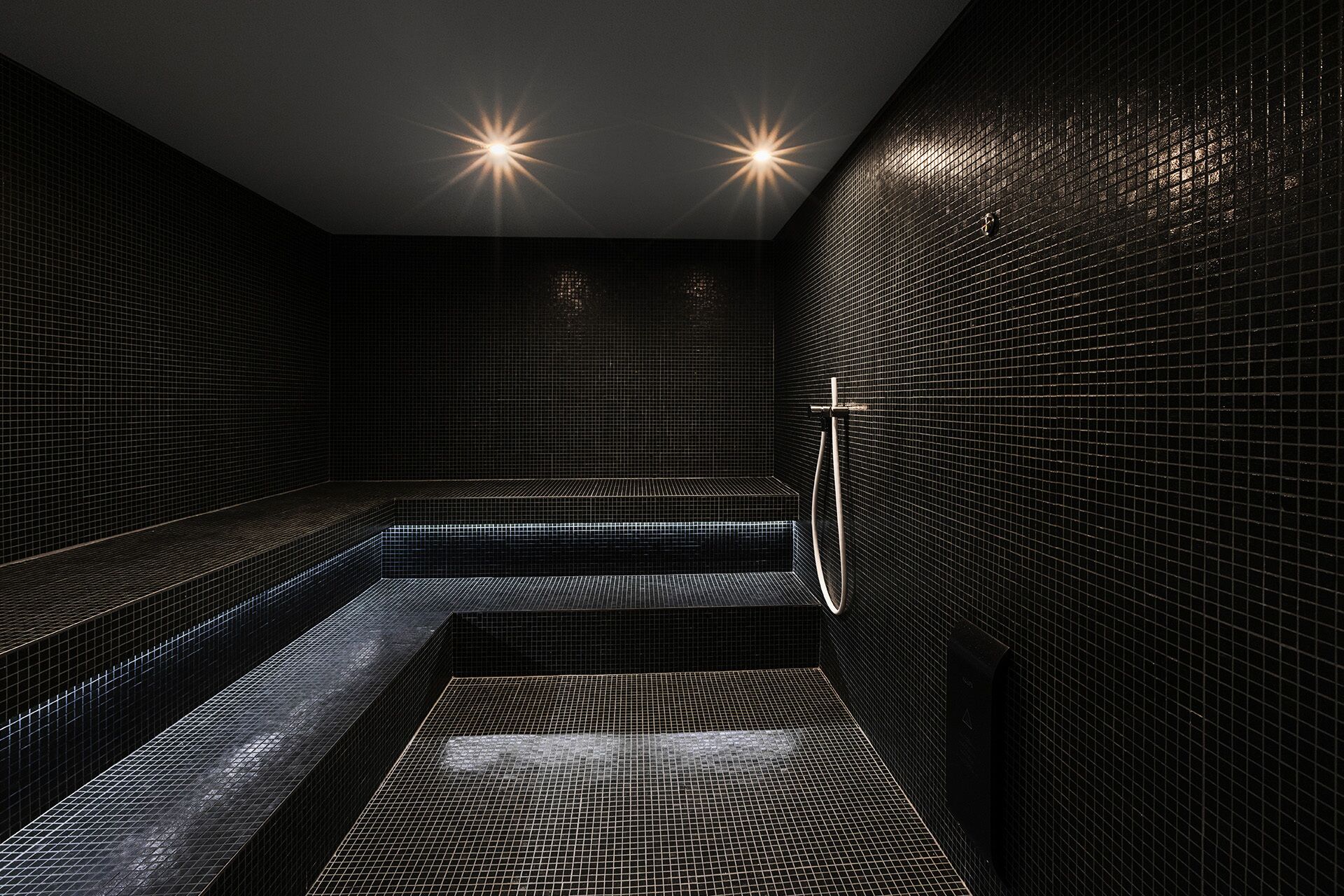 A modern, dimly lit spa room with black tiled walls and a central bench.
