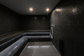 A modern, dimly lit spa room with black tiled walls and a central bench.