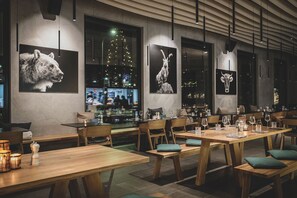 A restaurant interior with wooden tables and chairs, black and white animal-themed wall art, and a city view through large windows.