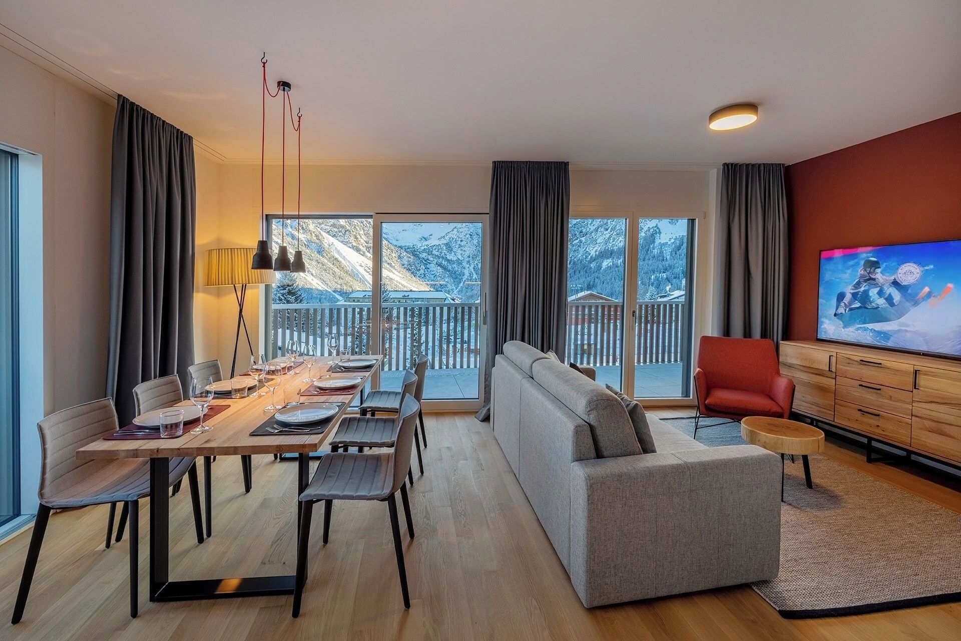 A modern living room with a dining area, a sofa, a television, and a view of snowy mountains.