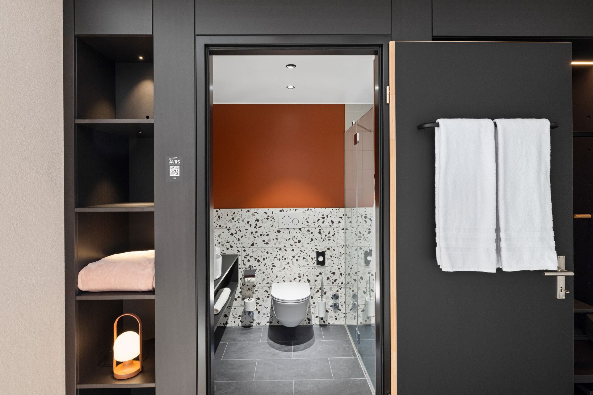 Une salle de bain moderne équipée de toilettes, d’un lavabo et d’étagères. On y trouve également un porte-serviettes avec deux serviettes.