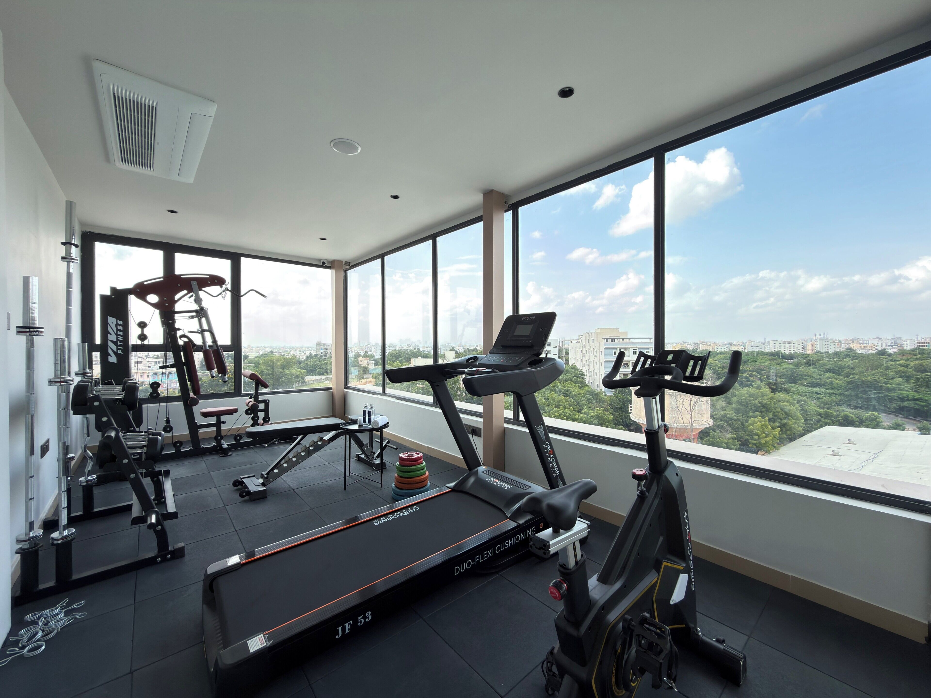 A gym with a treadmill, elliptical, and weight machines, large windows with a city view.