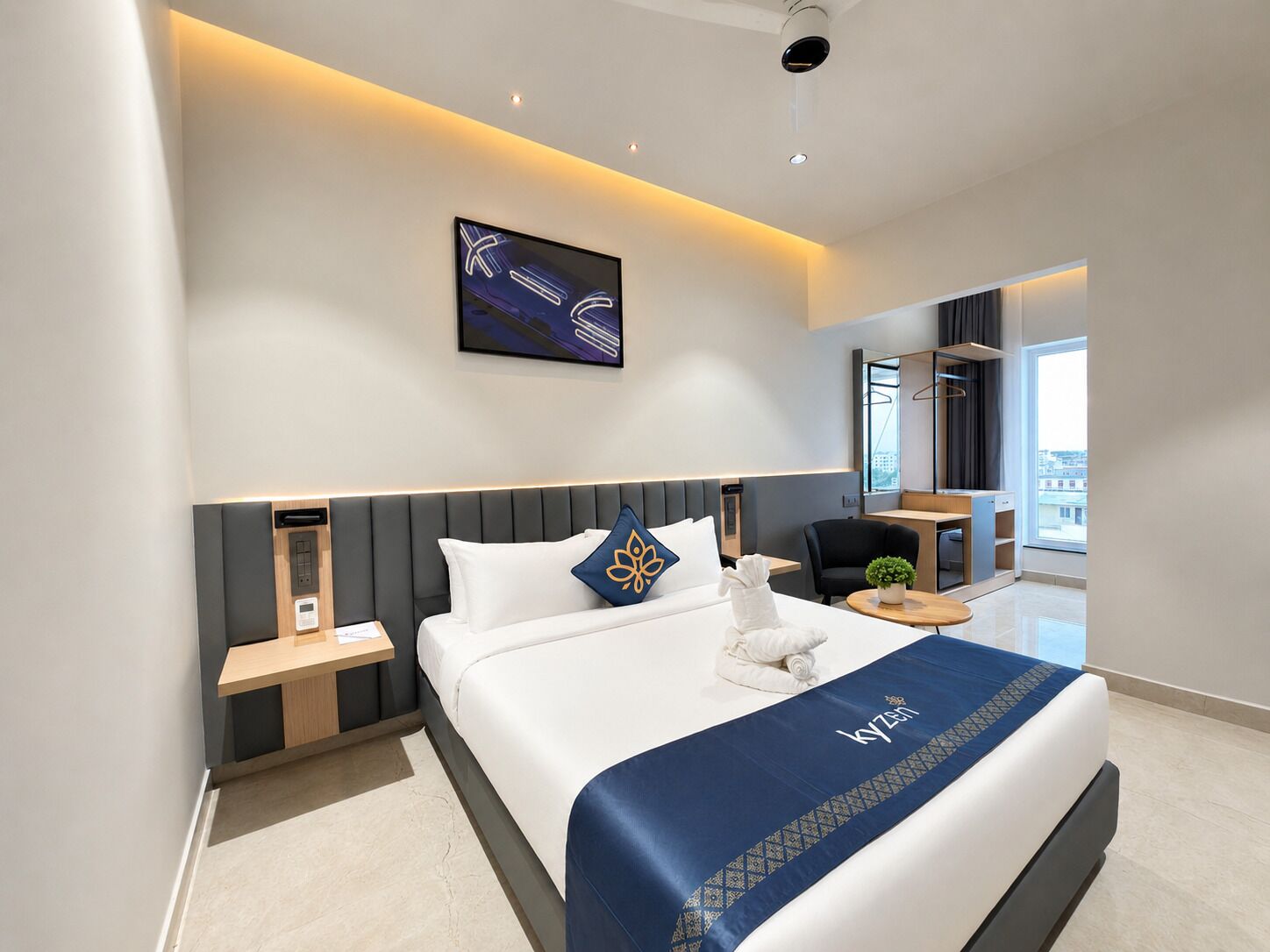 A modern hotel room with a large bed, a bedside table, a chair, a small table, a potted plant, and a city view.