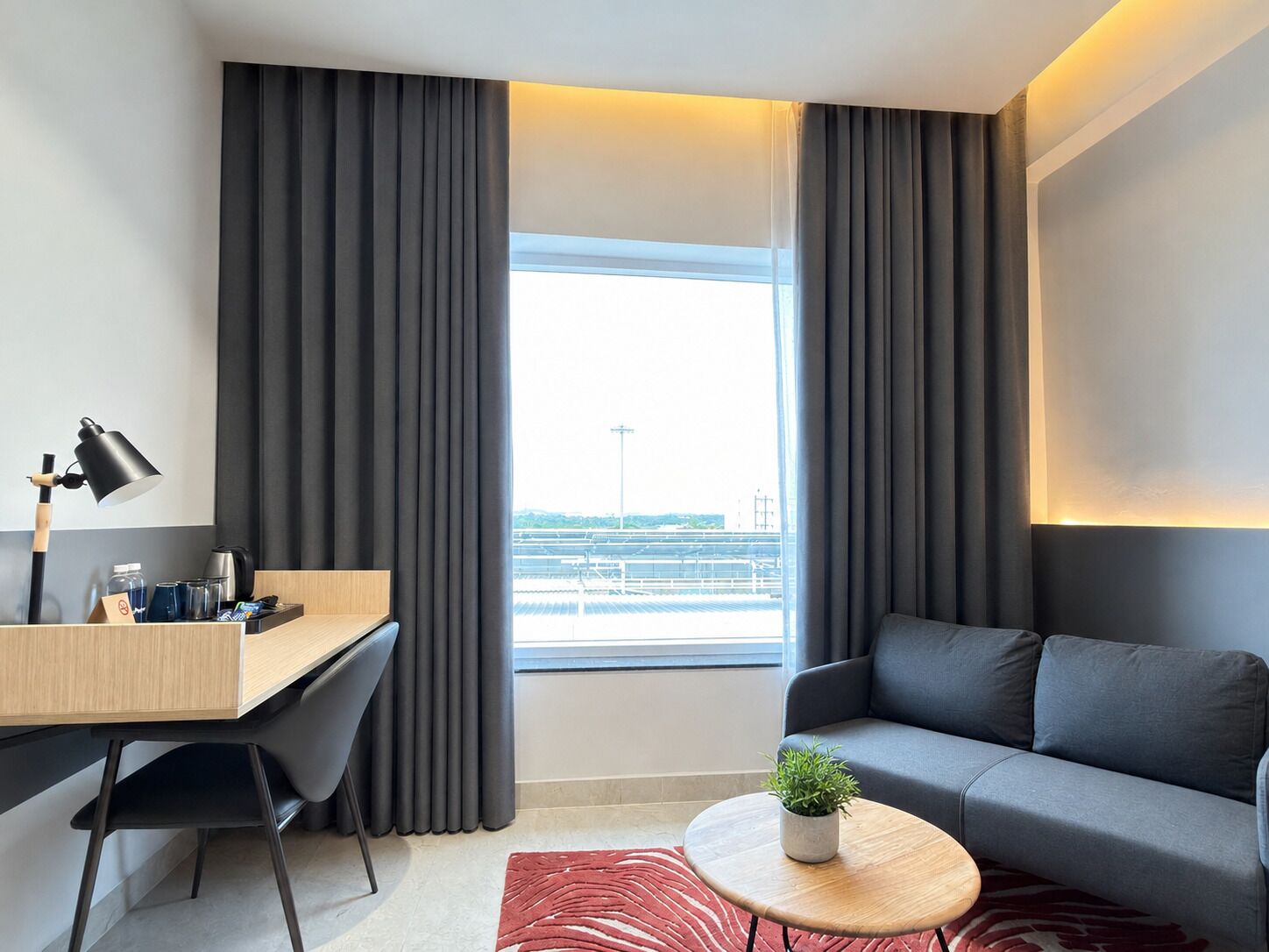 A modern hotel room with a desk, a sofa, a small table with a plant, and a view of the cityscape.