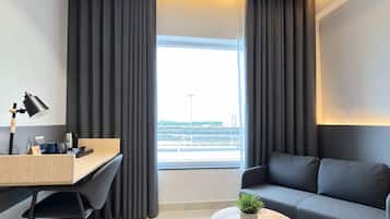 A modern hotel room with a desk, a sofa, a small table with a plant, and a view of the cityscape.