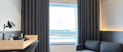A modern hotel room with a desk, a sofa, a small table with a plant, and a view of the cityscape.