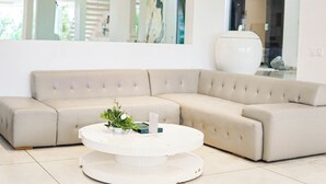 Een beige hoekbank met een witte ronde salontafel ervoor, een vaas met bloemen, en een grote witte vaas op een tafel in de achtergrond.