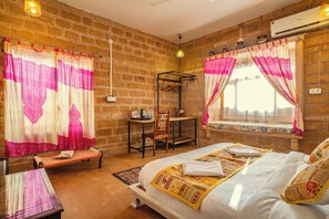 A room with a brick wall, a bed with a colorful quilt, a wooden chair, a small table with a kettle, and a window with curtains.