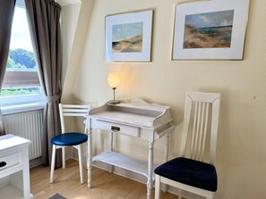 A room with a white desk, two chairs, a lamp, and two framed paintings on the wall. - Süderstraße 29a, Apt. 02 - 3-room upper-floor vacation apartment with a south-facing balcony for (Sylt-Ost)