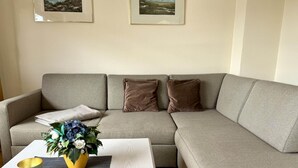 A living room with a grey sofa, two cushions, a small table with a vase of flowers, and two framed pictures on the wall. - Süderstraße 29a, Apt. 02 - 3-room upper-floor vacation apartment with a south-facing balcony for (Sylt-Ost)