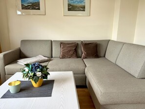 A living room with a grey sofa, two cushions, a small table with a vase of flowers, and two framed pictures on the wall. - Süderstraße 29a, Apt. 02 - 3-room upper-floor vacation apartment with a south-facing balcony for (Sylt-Ost)