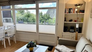 A cozy living room with a sofa, a coffee table with a plant, a bookshelf, and a view of the outdoors through large windows. - Süderstraße 29a, Apt. 02 - 3-room upper-floor vacation apartment with a south-facing balcony for (Sylt-Ost)