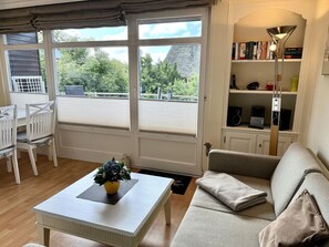 A cozy living room with a sofa, a coffee table with a plant, a bookshelf, and a view of the outdoors through large windows. - Süderstraße 29a, Apt. 02 - 3-room upper-floor vacation apartment with a south-facing balcony for (Sylt-Ost)